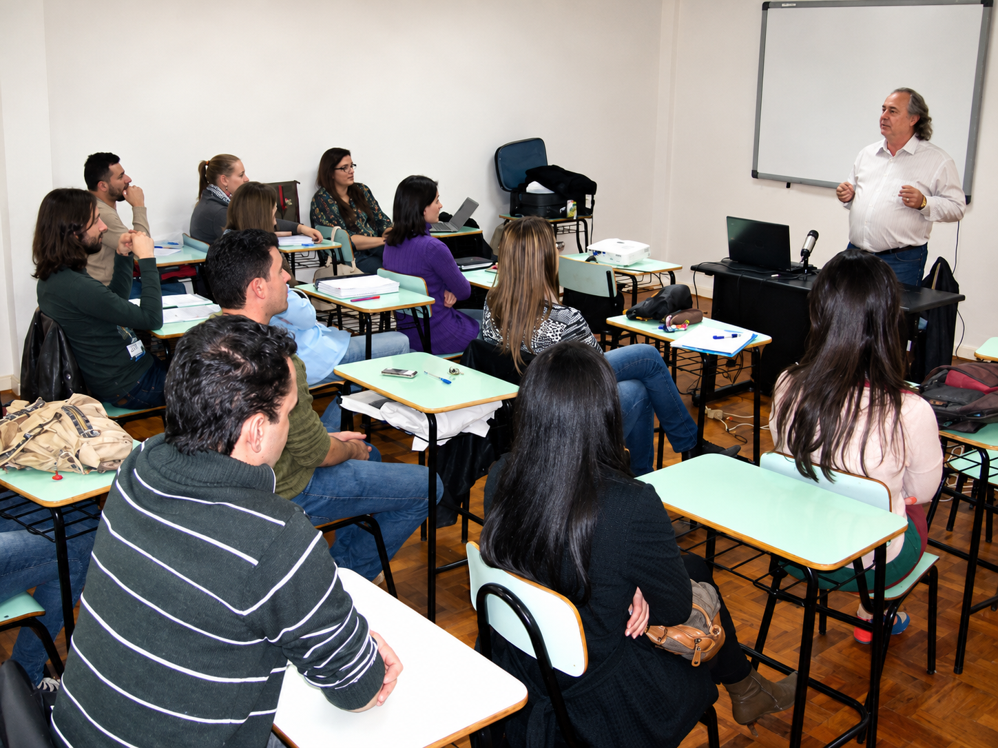 Fotografia de uma sala de aula: um professor está em pé, próximo ao quadro, diante de participantes sentados em carteiras.