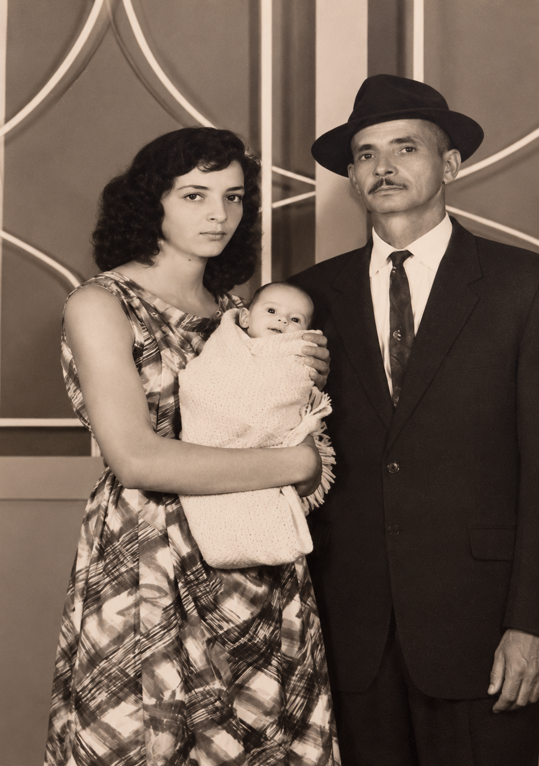 Fotografia antiga em tons sépia: uma mulher segura um bebê nos braços, ao lado de um homem de terno e gravata.